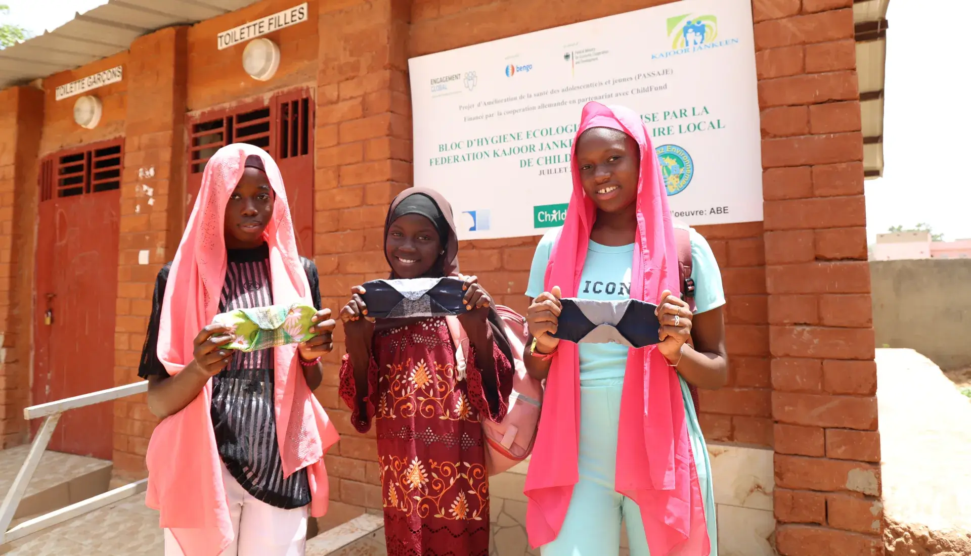 Fatou, 13 ans, Zainab, 14 ans, et Mame, 14 ans, tenant des serviettes hygiéniques réutilisables devant le bloc de toilettes et d’hygiène menstruelle de leur école.